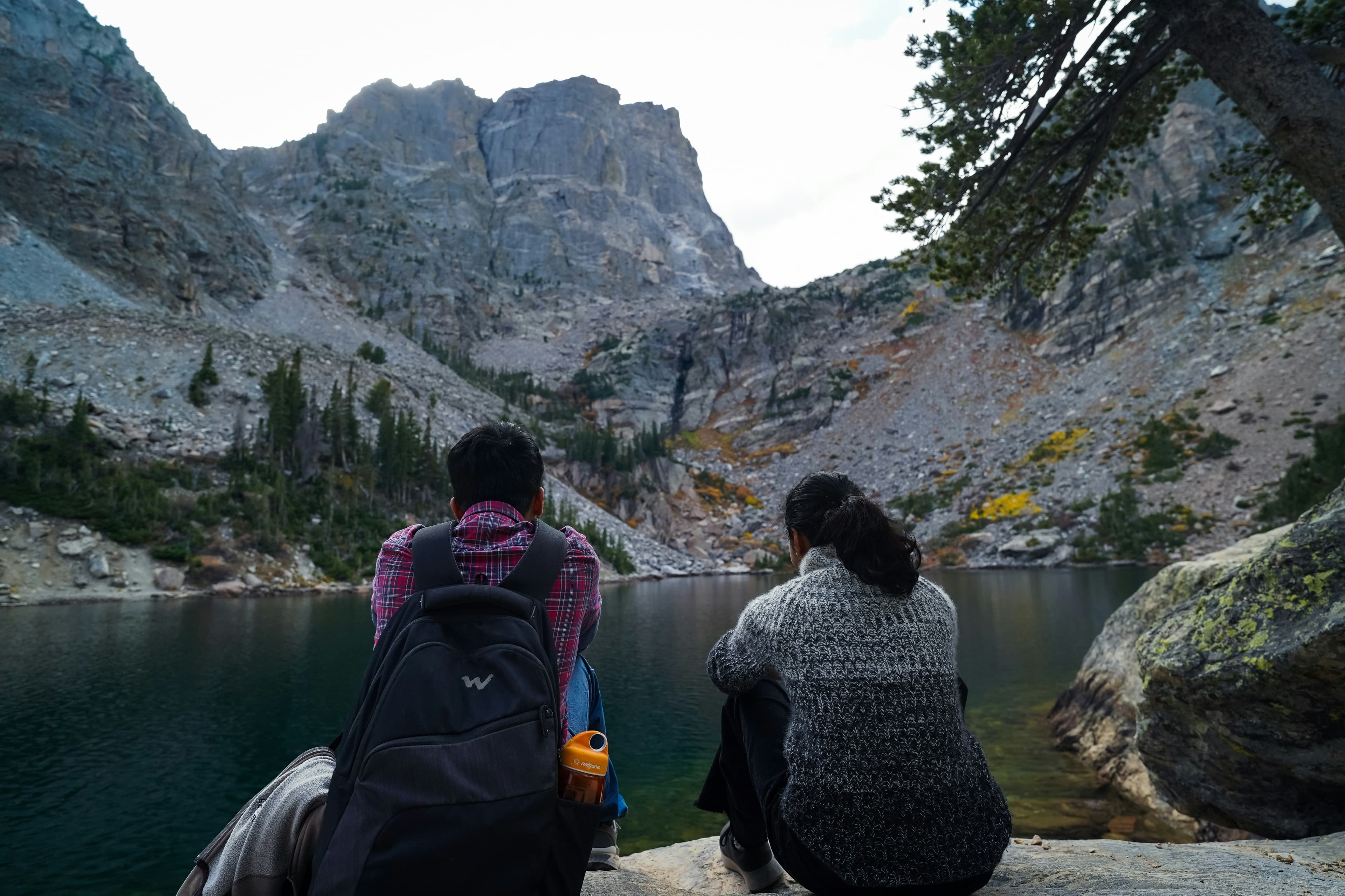 Two people sitting beside a mountain lake beneath rocky cliffs.